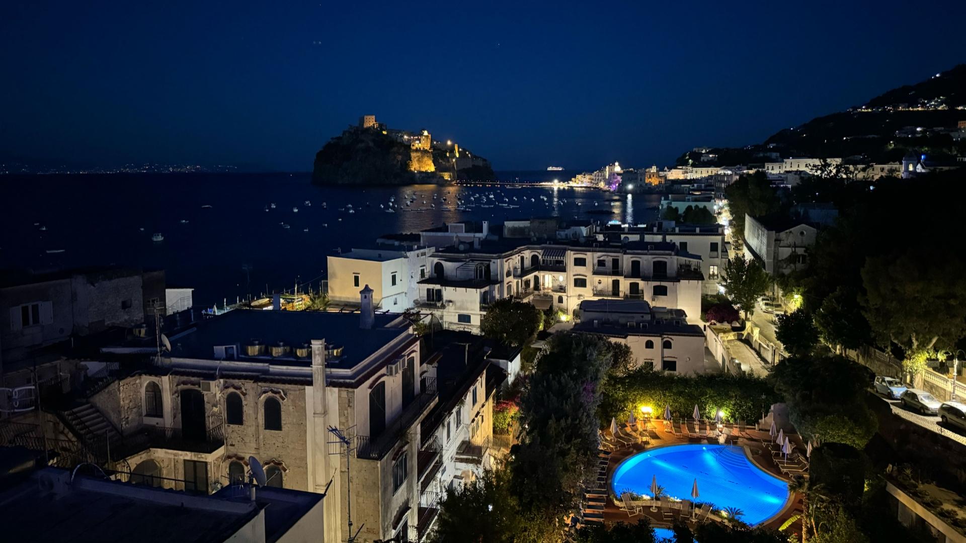 Vista notturna suggestiva su mare, castello illuminato e piscina blu, atmosfera magica e serena.