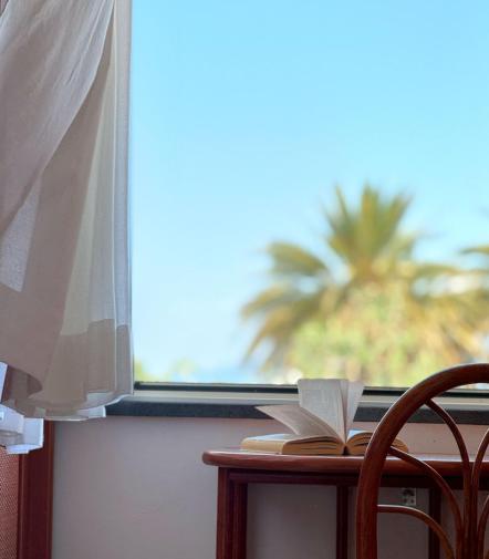 Una finestra aperta, tende leggere, libro sfogliato e palme sotto un cielo sereno.