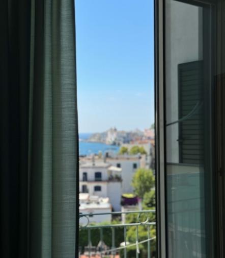 Vista da una finestra aperta su un paesaggio costiero, cielo azzurro e case bianche.