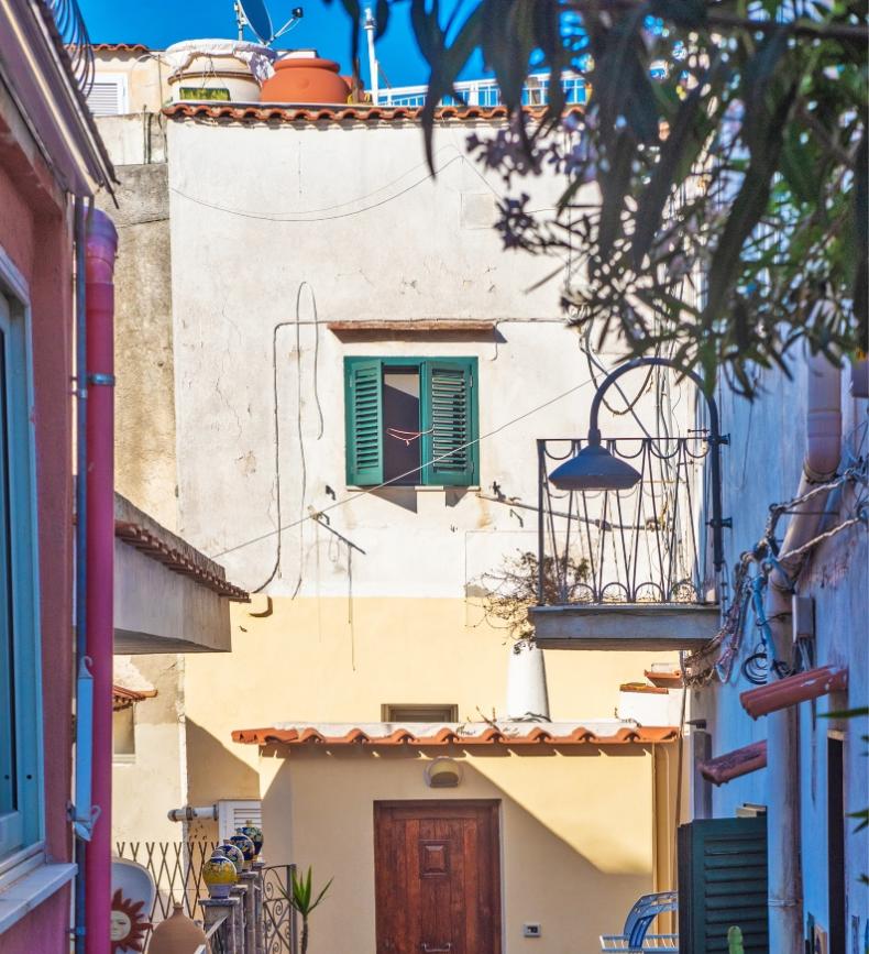 Vicolo pittoresco con case colorate, piante rigogliose e atmosfera mediterranea sotto un cielo azzurro.
