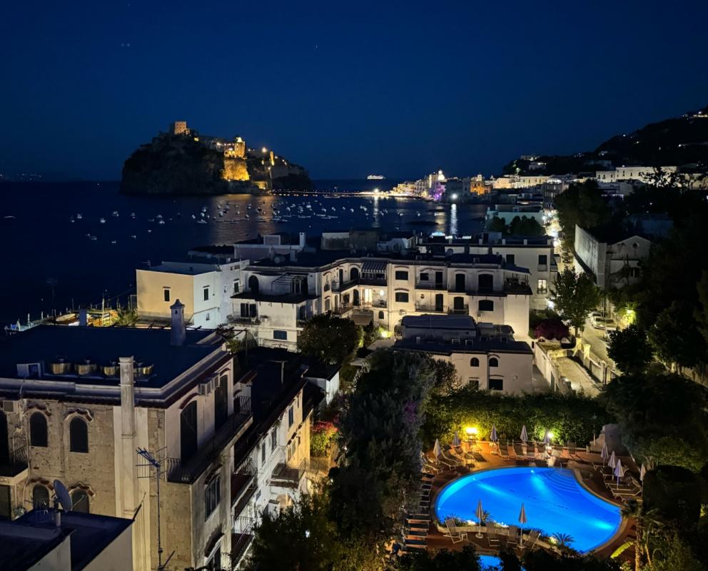 Vista notturna suggestiva su mare, castello illuminato e piscina blu, atmosfera magica e serena.